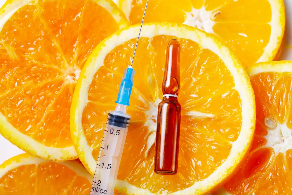 Image of fresh orange slices arranged in the background with a syringe and a small amber vial placed on top, symbolizing the concept of vitamin C injections or supplements.